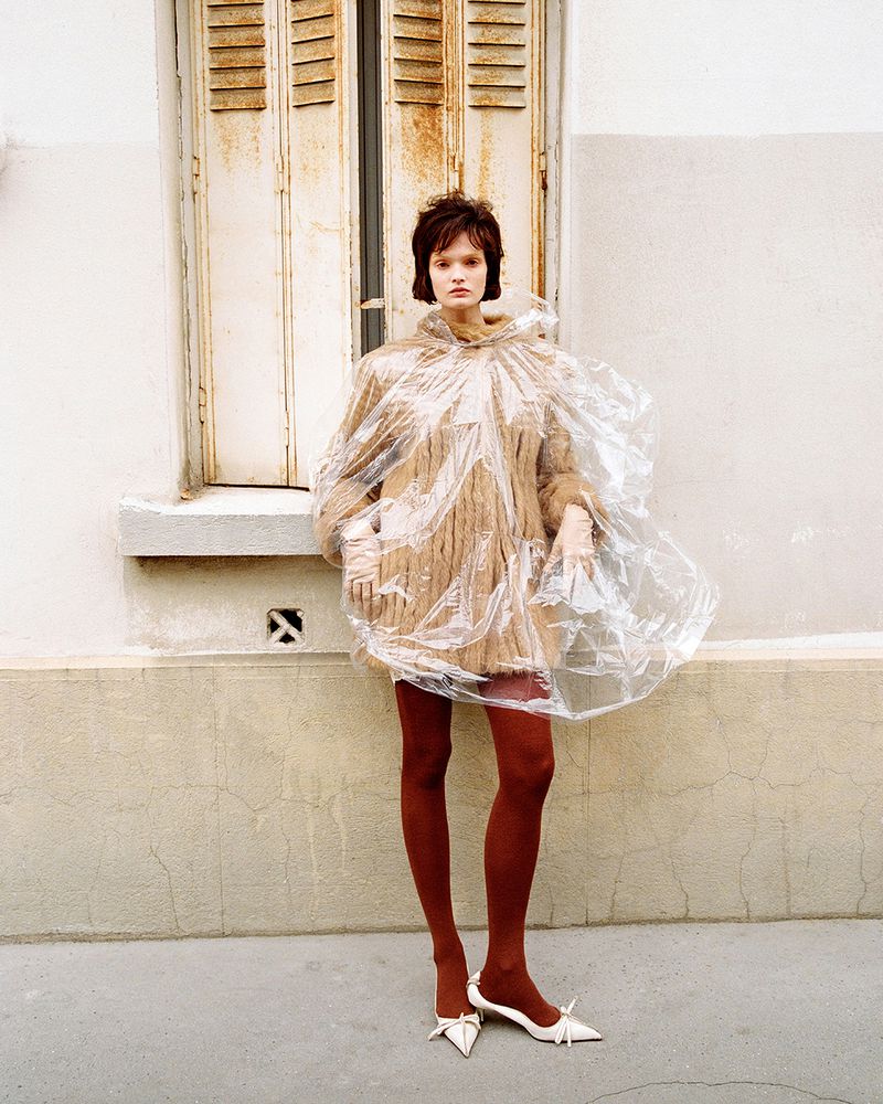 Woman posing in a plastic coat in front of a window with shutters, wearing a dress, tights, and heels for a fashion shot outdoors.