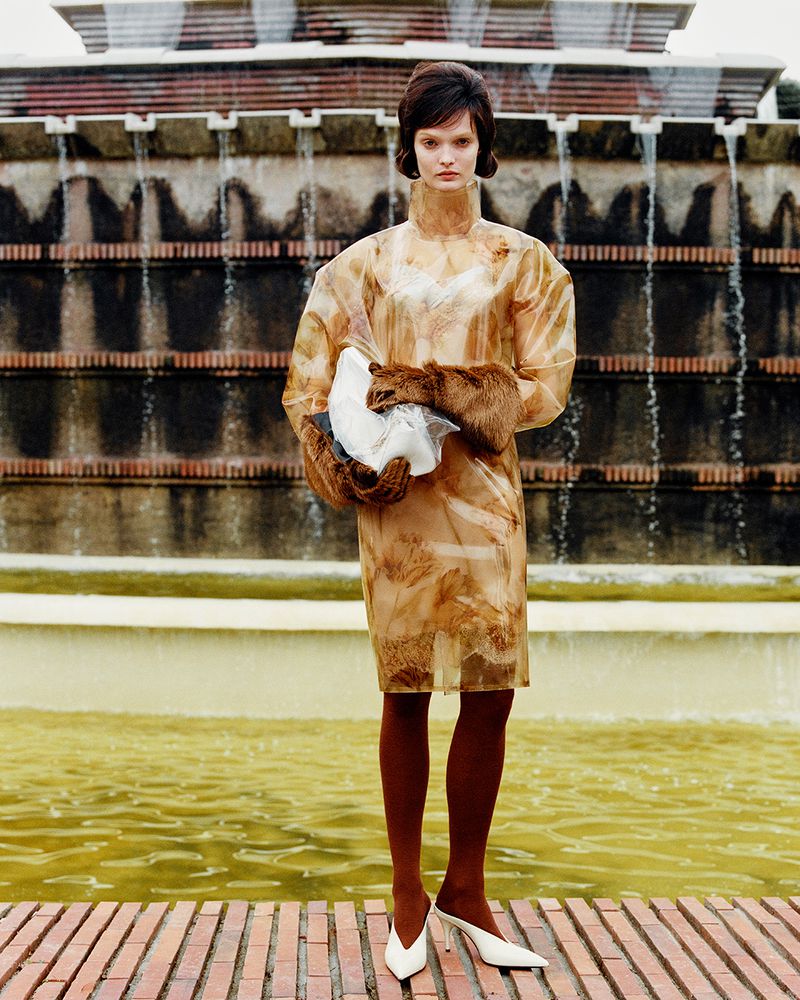 Fashion shot featuring a brunette adult woman in a patterned dress with white heels, posed in front of a tiered fountain.
