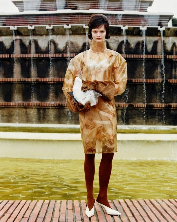 Fashion shot featuring a brunette adult woman in a patterned dress with white heels, posed in front of a tiered fountain.