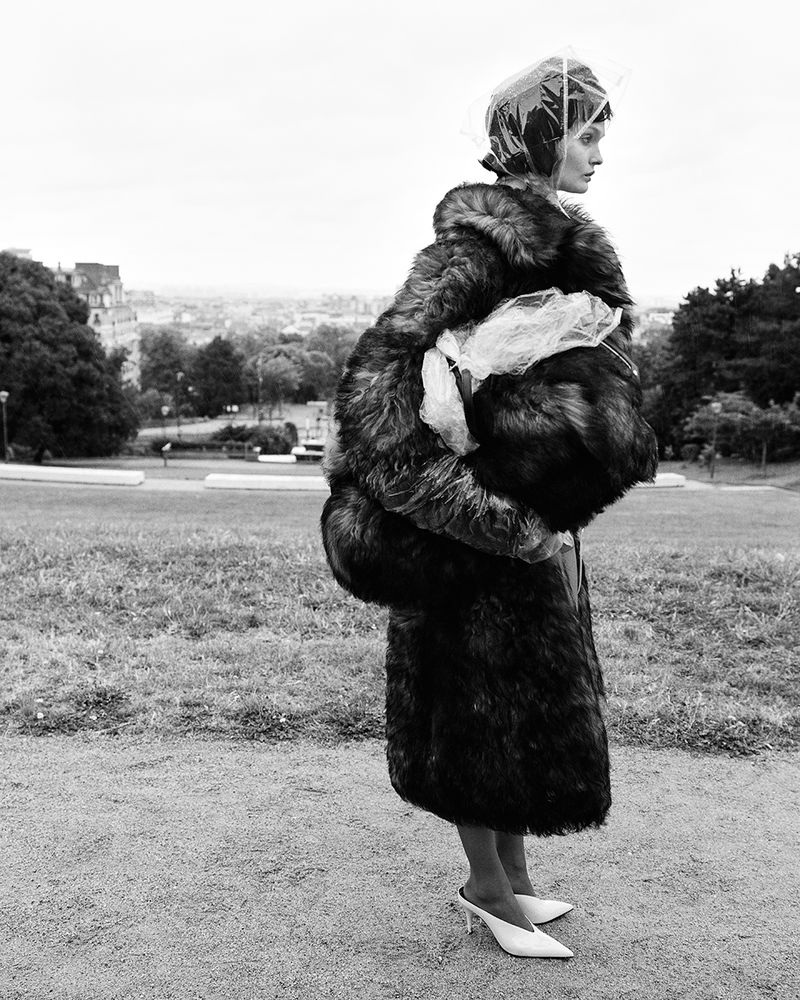 Monochrome portrait of a stylish woman in a fur coat and white heels, posing on a hill with a cityscape in the background.