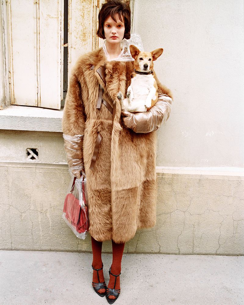 Model Lulu Tenney in a long camel fur coat holding a small tan and white dog against an exterior stucco wall.