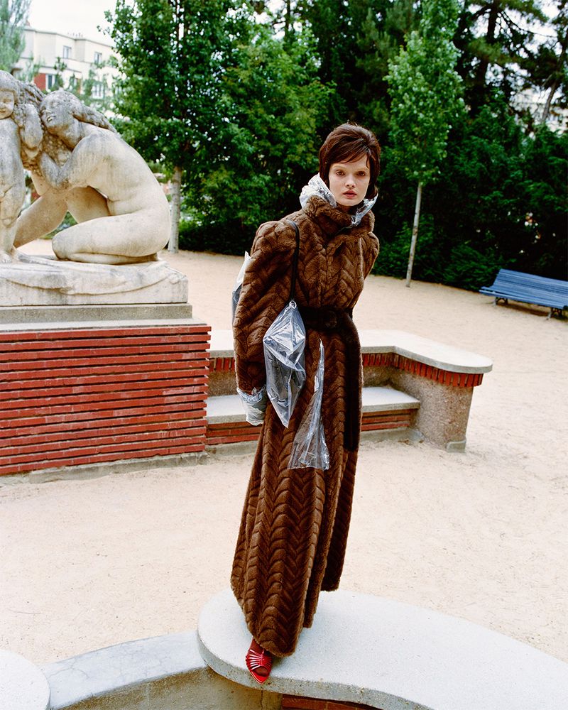 Woman in a brown chevron maxi coat with red shoes poses near a stone sculpture in an outdoor park-like setting with trees and green space.