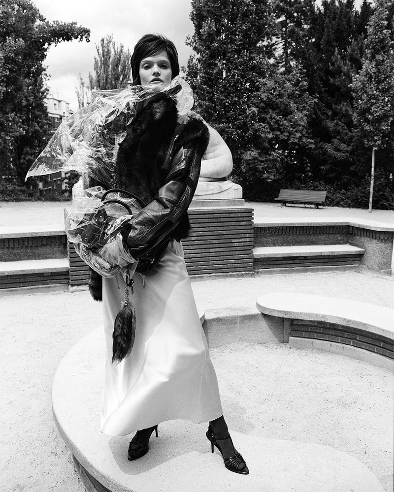 A monochrome outdoor fashion portrait of an adult woman with short dark hair, wearing a leather jacket and long dress in a park setting.