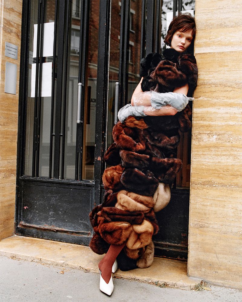 Lulu Tenney leans against a building doorway wearing a sculptural tiered brown and black fur dress with white pointed mules.