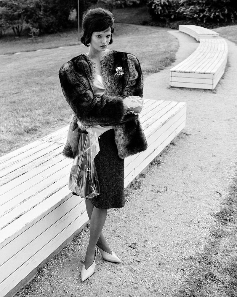Black and white image of a woman in a fur coat standing next to a bench, with trees in the background.