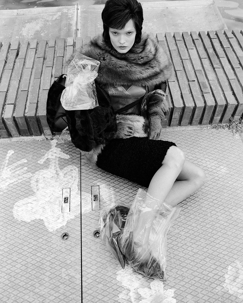 Black and white fashion photo of a young woman with dark hair wearing a fur shawl and skirt, sitting on a concrete platform.