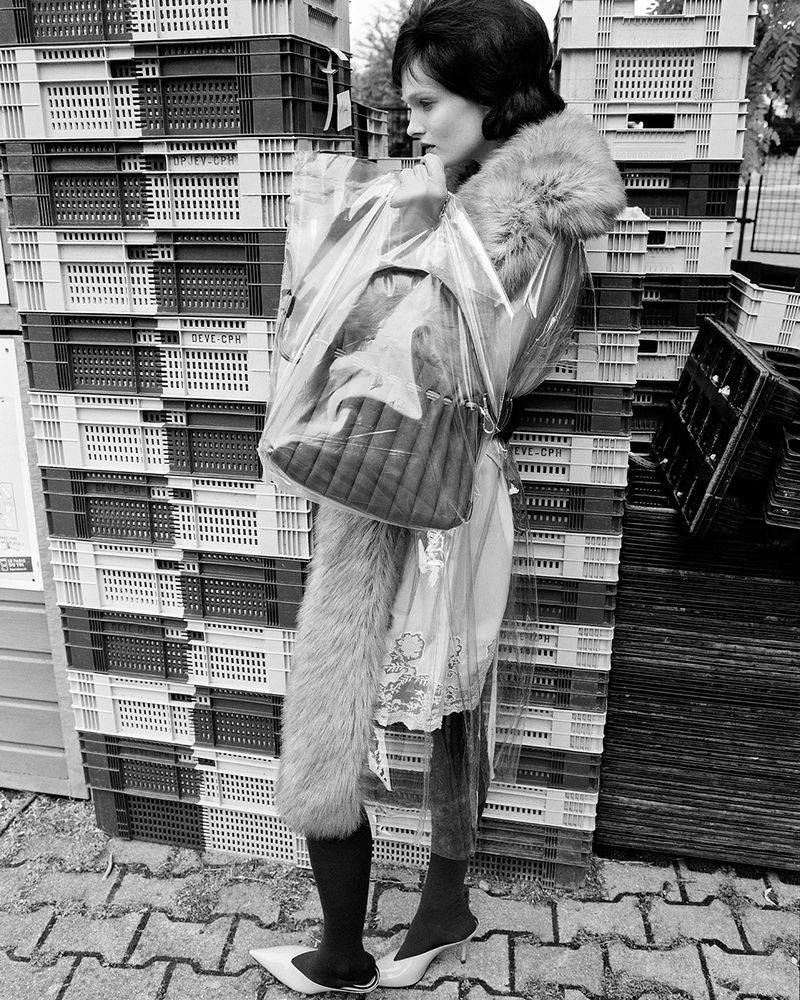 Black and white image of a stylish adult woman posing outdoors with a fur-lined coat, handbag in plastic, and patterned background.