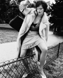 Model Lulu Tenney wears an oversized plush coat and satin skirt while straddling a low wire park fence in a black and white editorial.