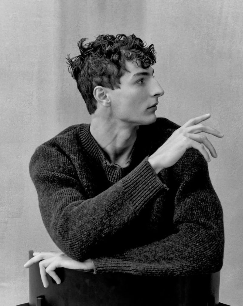 Monochromatic portrait of a young adult male with curly hair wearing a knit sweater, seated and looking off to the side.
