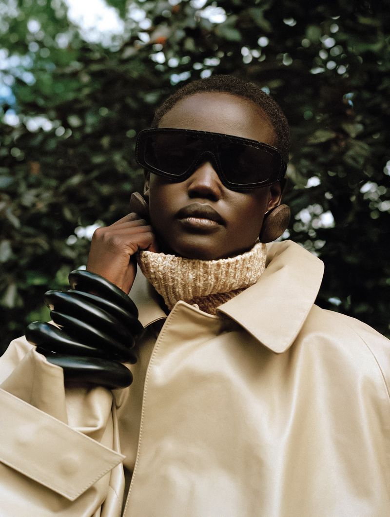 Fashion portrait of a Black woman with sunglasses, a turtleneck sweater, a trench coat, and oversized bracelets in a leafy setting.