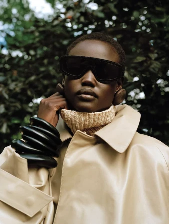 Fashion portrait of a Black woman with sunglasses, a turtleneck sweater, a trench coat, and oversized bracelets in a leafy setting.