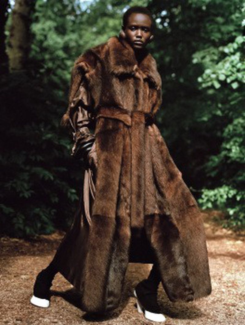 A dark-skinned model in a full-length mink coat poses in a forest setting with greenery visible behind her in a fashion-forward editorial style.