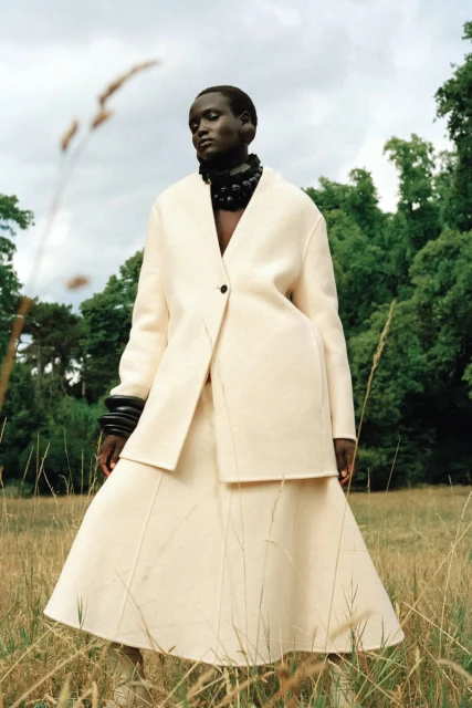 Woman wearing cream-colored coat and skirt in a grassy field with trees in the background, fashion photography, outdoor setting.