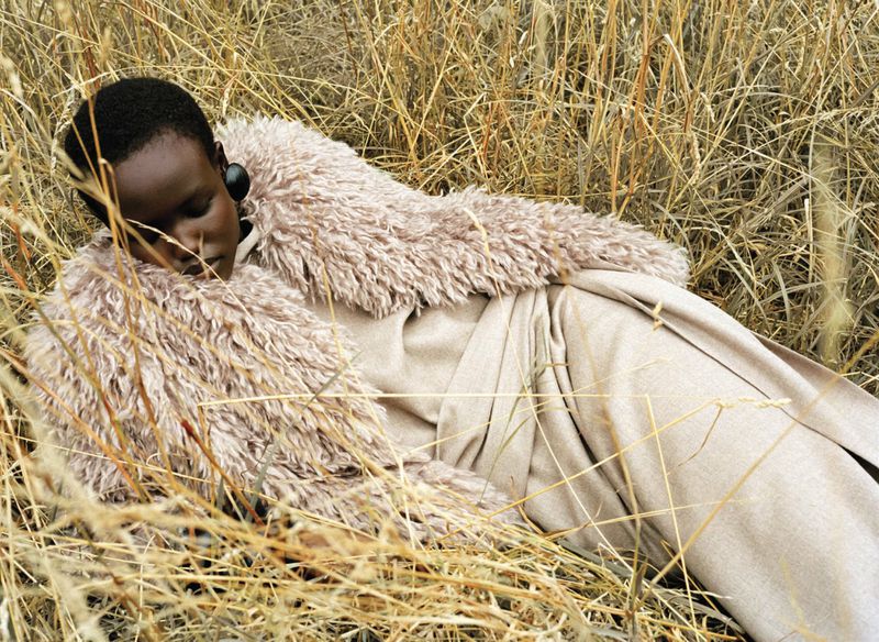 Woman in a fur-collared coat lying in tall, dry grass in a fashion shot featuring an earth-tone color palette and natural outdoor setting.