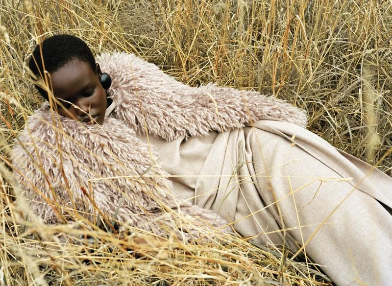 Woman in a fur-collared coat lying in tall, dry grass in a fashion shot featuring an earth-tone color palette and natural outdoor setting.