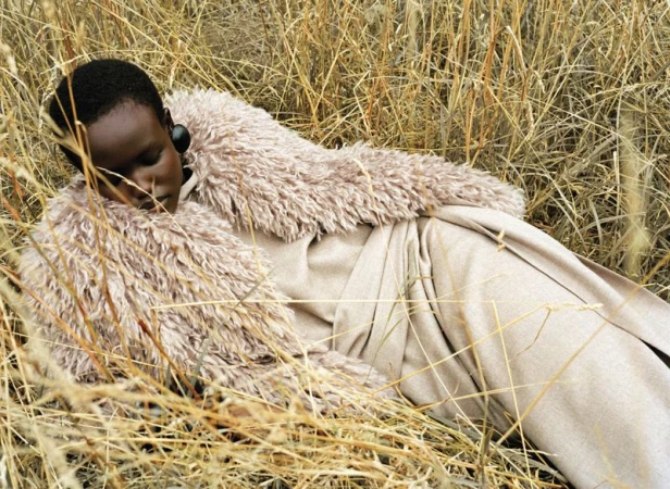 Woman in a fur-collared coat lying in tall, dry grass in a fashion shot featuring an earth-tone color palette and natural outdoor setting.