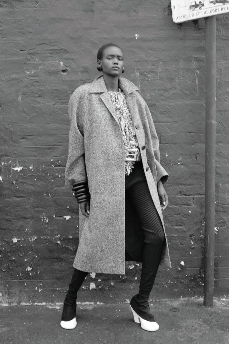 Black and white fashion shot of a model in a long coat, patterned shirt, leggings, and black and white heeled boots against a brick wall.