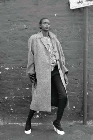Black and white fashion shot of a model in a long coat, patterned shirt, leggings, and black and white heeled boots against a brick wall.