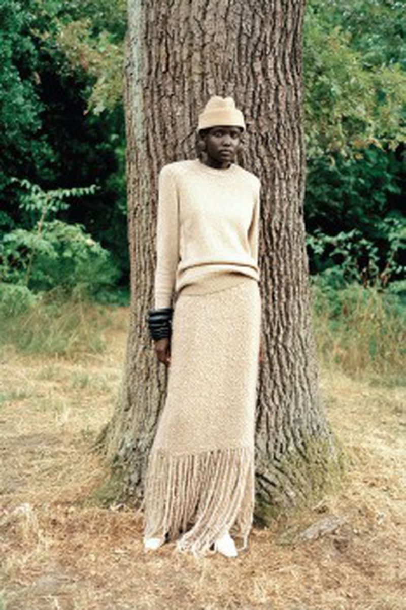 Woman in beige sweater and fringed skirt poses near a tree in a forest-like setting, wearing matching hat and thick black cuff bracelets.