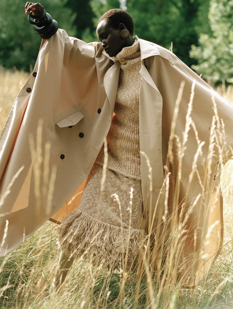 Woman in beige trench coat stands in a field of tall grass on a sunny day, fashionable pose with outspread arms, warm tones.