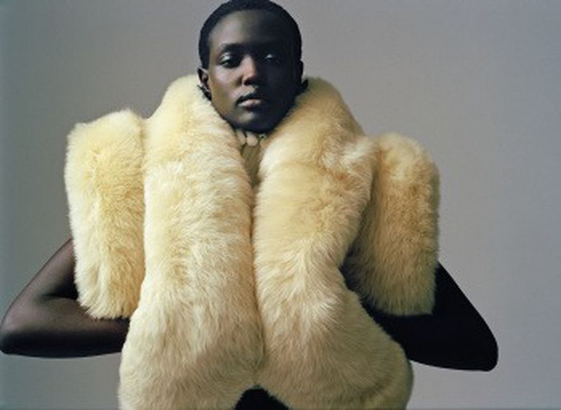 Fashion portrait of an adult African male with dark skin, wearing a cream-colored faux fur stole, posing in front of a grey background.