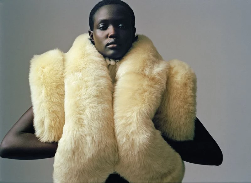 Portrait of an adult woman with dark skin, neutral expression, and bare arms, posing in a studio with a pale yellow faux fur stole.