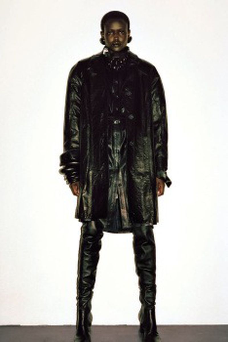 Fashionable young adult stands confidently in a black coat and thigh-high boots against a bright background in a studio setting.