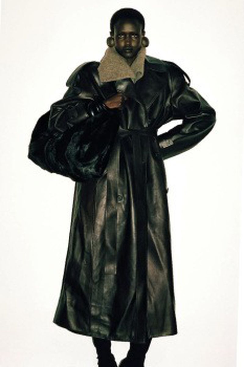Model Yar Aguer wears an oversized black leather trench coat with large disc earrings against a stark white studio backdrop.