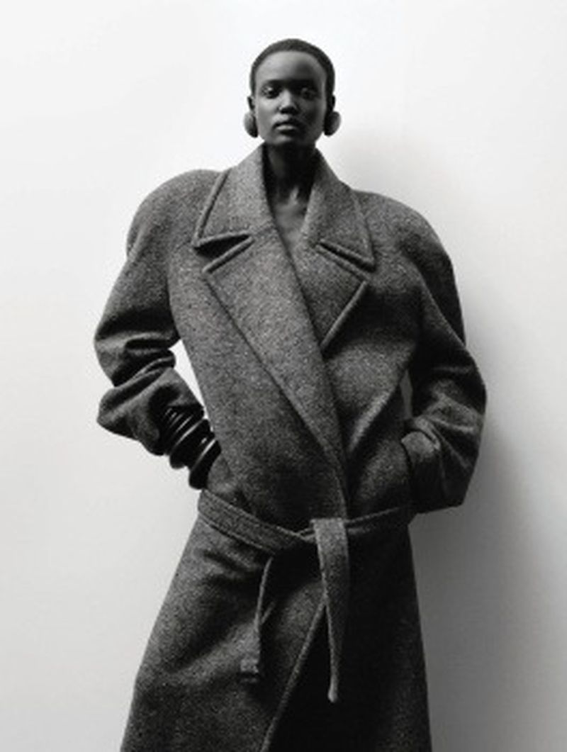 Elegant woman in a grey wool coat with a tie belt posing against a simple white background in a studio fashion shot.