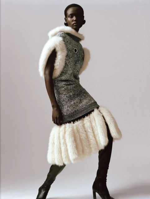 Fashion editorial shot of a dark-skinned woman in a grey dress trimmed with white faux fur, posing against a pale grey backdrop.