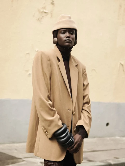 Fashionable woman in a tan blazer and hat posing against a textured wall, showcasing 1980s-inspired style with neutral tones and bold accessories.