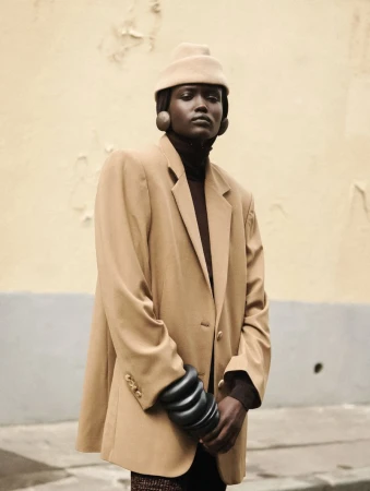 Fashionable woman in a tan blazer and hat posing against a textured wall, showcasing 1980s-inspired style with neutral tones and bold accessories.