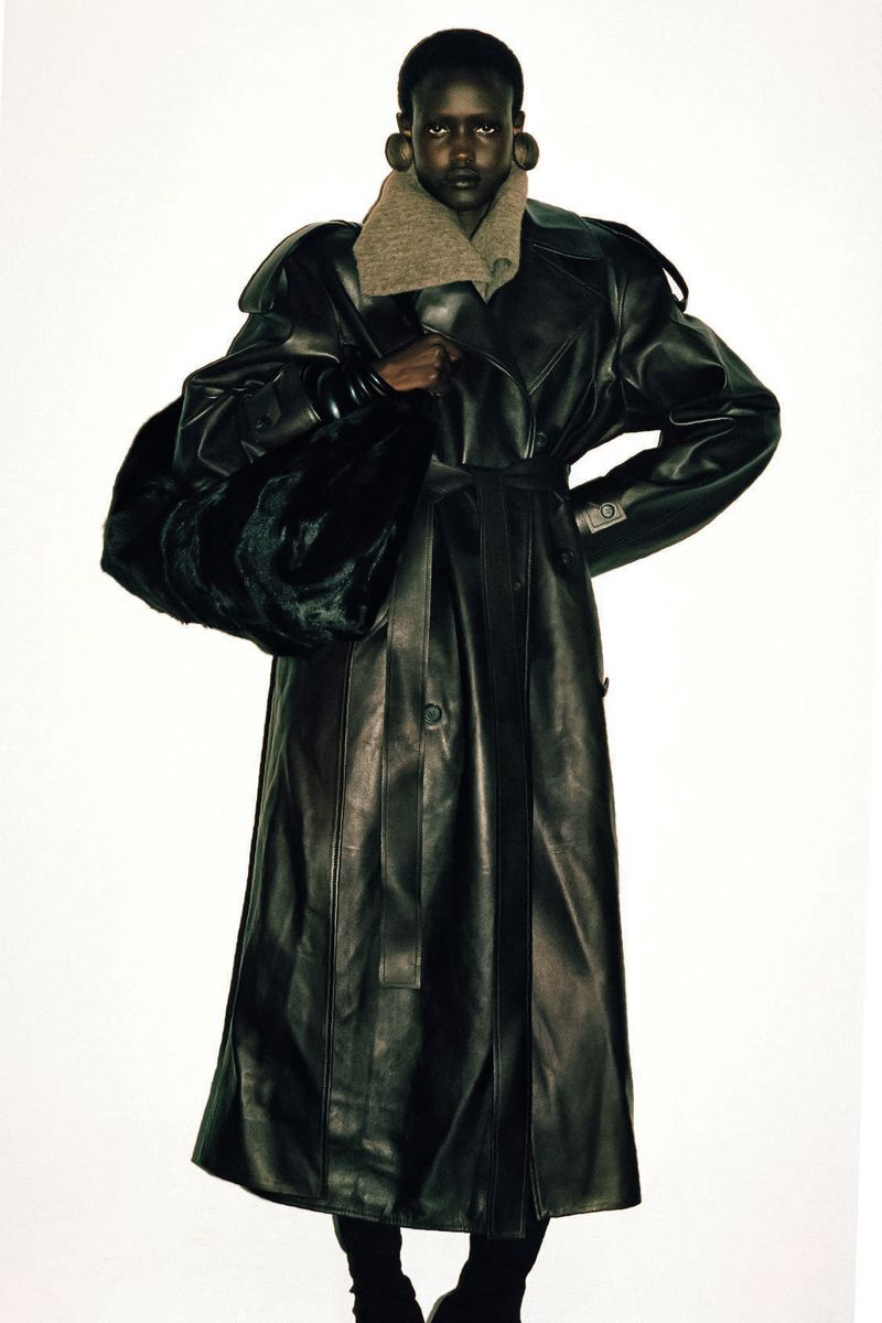 Fashionable dark-skinned model poses in a studio wearing a long leather trench coat and carrying a large fur bag against a white backdrop.