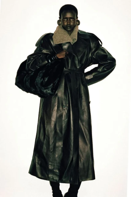 Fashionable dark-skinned model poses in a studio wearing a long leather trench coat and carrying a large fur bag against a white backdrop.