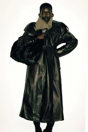 Fashionable dark-skinned model poses in a studio wearing a long leather trench coat and carrying a large fur bag against a white backdrop.