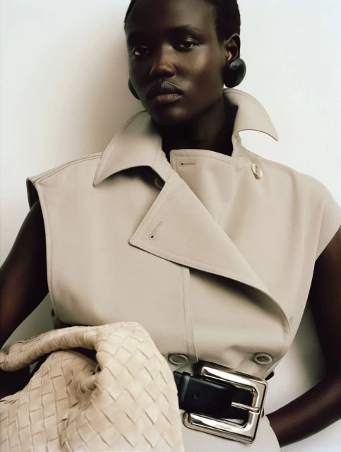 Close-up of a Black woman posing indoors, wearing a beige sleeveless trench coat and accessorized with dark earrings and a large woven handbag.