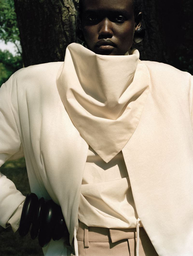 Close-up fashion portrait of a black woman wearing an oversized cream jacket in a wooded outdoor setting.