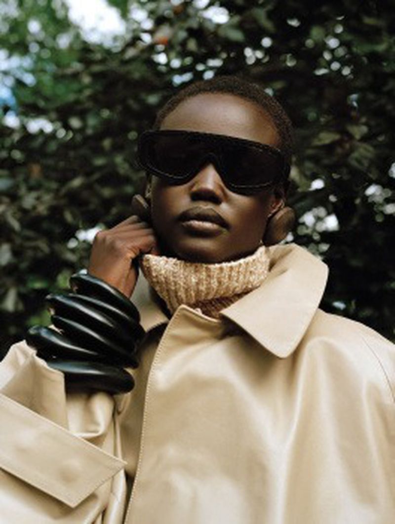 Close-up studio shot of a dark-skinned adult female model with large sunglasses and black bracelets against an out-of-focus green background.