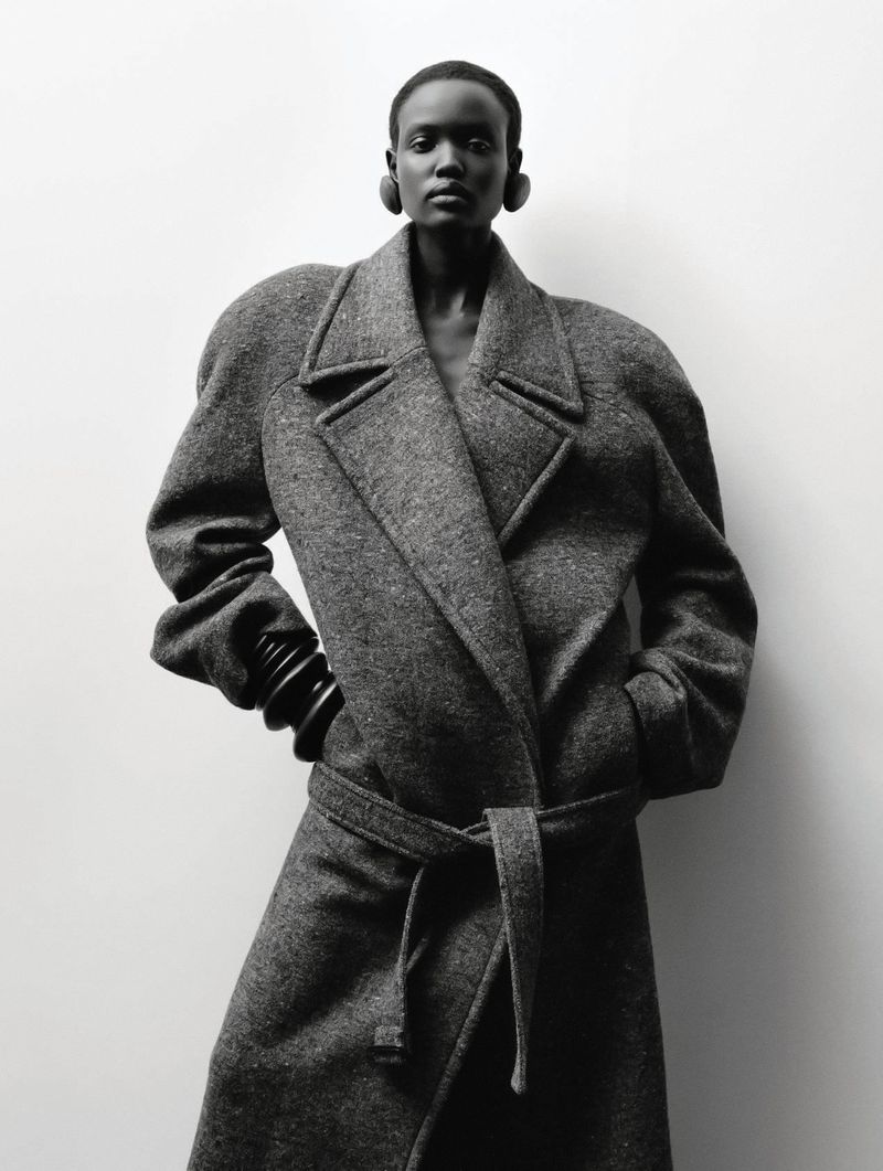 Fashion shot of a Black woman in a grey wool coat with a tie belt, wearing black hoop earrings and bangle bracelets, against a neutral backdrop.