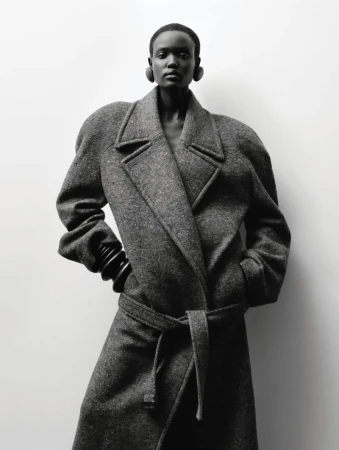 Fashion shot of a Black woman in a grey wool coat with a tie belt, wearing black hoop earrings and bangle bracelets, against a neutral backdrop.
