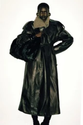 Model Yar Aguer wears an oversized black leather trench coat with large disc earrings against a stark white studio backdrop.