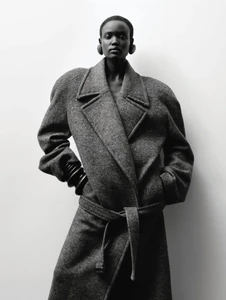 Model Yar Aguer in an oversized belted wool coat and sculptural earrings posing against a plain backdrop in black and white.