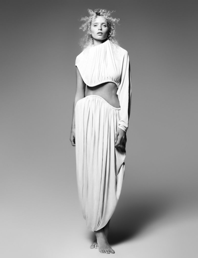 Black and white fashion portrait of a woman wearing a draped, one-sleeved white top and skirt, standing in a studio setting.