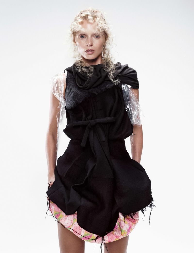 Stylish portrait of a blonde woman in a black dress with plastic accents, posing in a studio with soft lighting and a neutral background.
