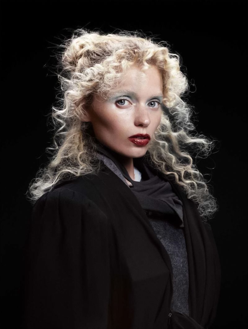 Portrait of a woman with curly blonde hair and dark lipstick wearing a black coat against a black background in a studio setting.