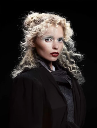 Portrait of a woman with curly blonde hair and dark lipstick wearing a black coat against a black background in a studio setting.