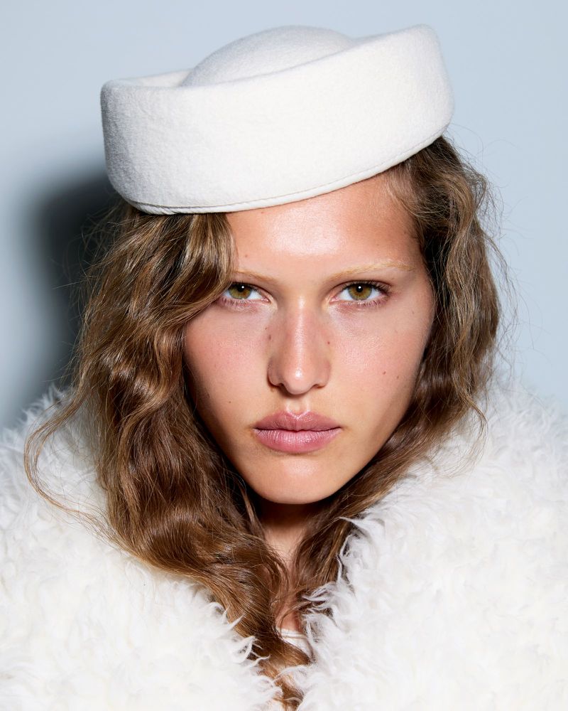 Close-up portrait of a fair-skinned woman with wavy brown hair wearing a white pillbox hat and fluffy white coat against light blue.