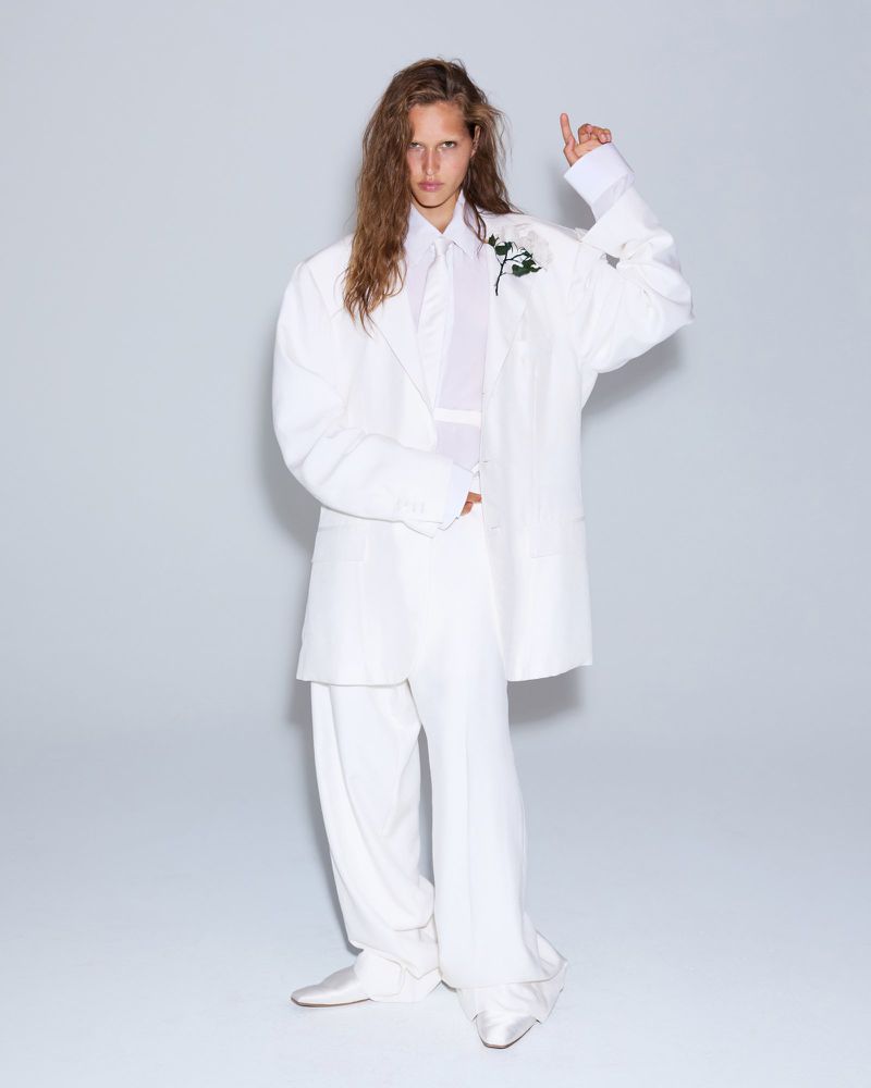 Person in oversized white suit with long hair poses in a full shot in front of a white background, pointing upward, wearing a floral corsage.