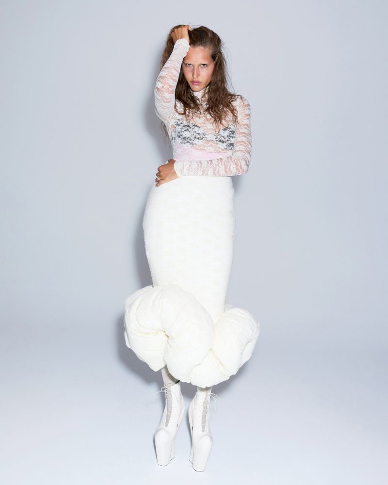 Fashionable woman strikes a pose in a white dress, lace top, and unique heeled shoes in a studio setting with even lighting.