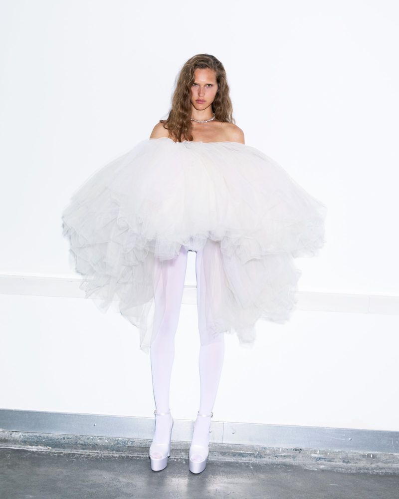 A woman wearing a large white tulle skirt and matching tights and platform heels stands against a white wall in a fashion-style photo.